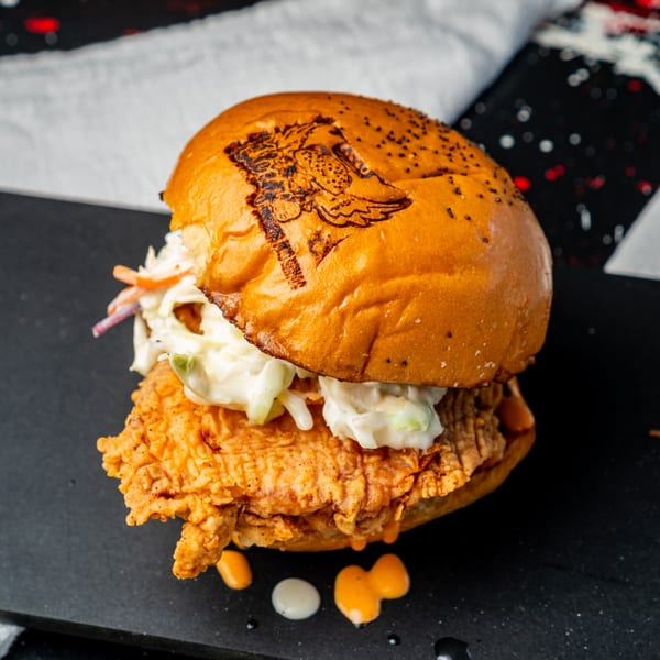 Little Angel Burgers & Wings chicken sandwich with coleslaw and branded bun on a dark surface.