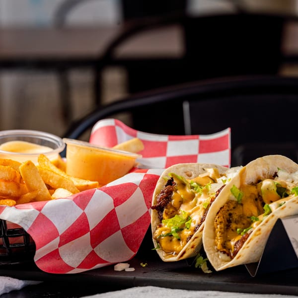 Two tacos with meat, cheese, and lettuce, next to a basket of fries and dipping sauce.